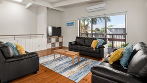 A modern living room with a wooden coffee table, two black leather sofas with yellow and blue cushions, a flat-screen TV, and a large window with a view of palm trees and a house. - Spacious 4-bed Home 5-min From the Beach (Melbourne)