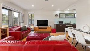 A modern living room with red leather sofas, a flat-screen TV, and a dining area with white chairs. - Spacious 4-bed Home 5-min From the Beach (Melbourne)
