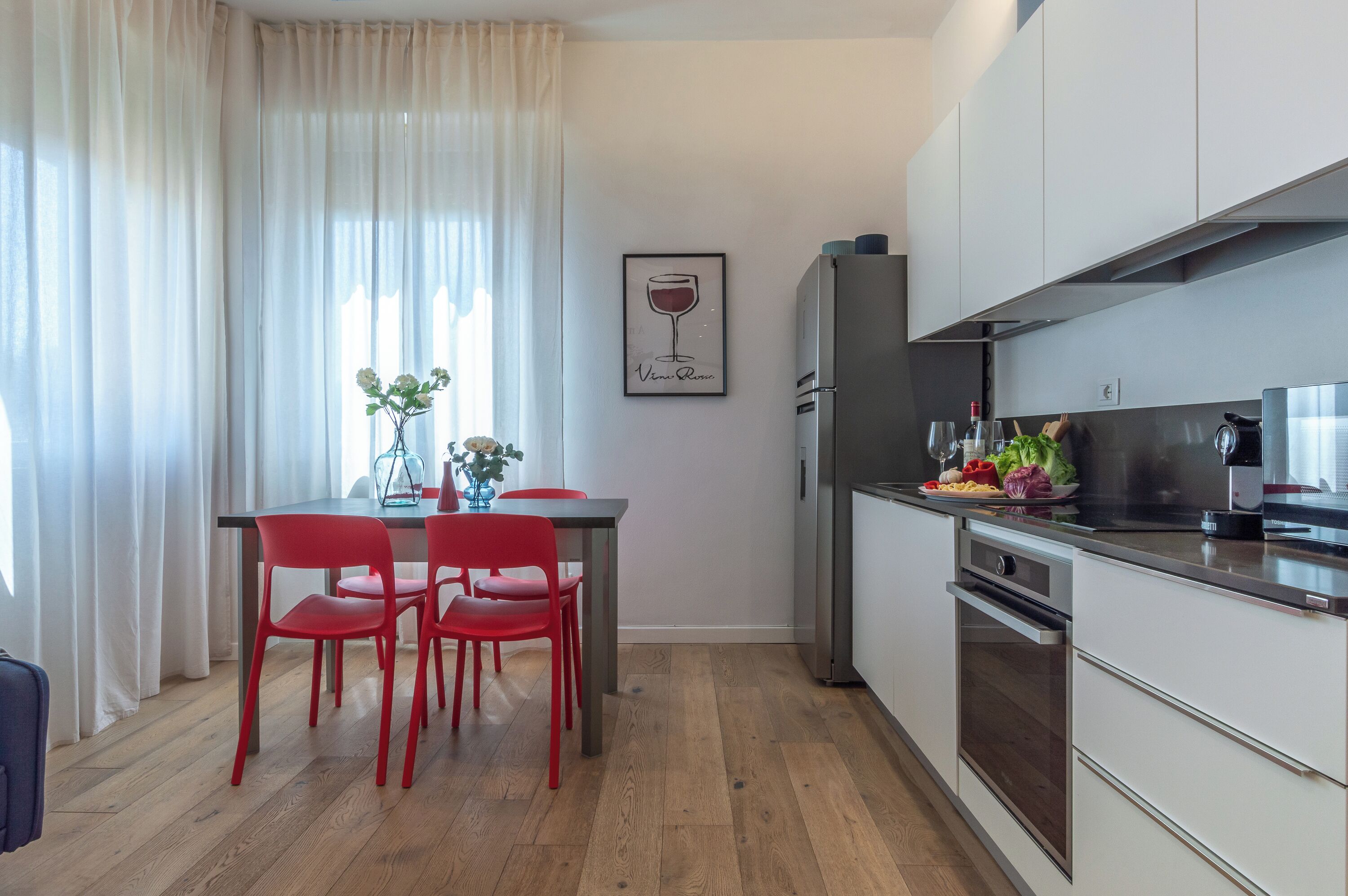 A modern kitchen with a dining area, featuring a table with red chairs, a refrigerator, and a stove.