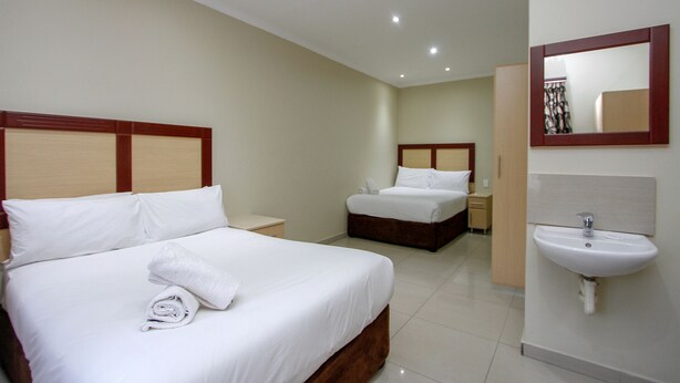 A hotel room with two beds, a bedside table, a nightstand, and a bathroom with a sink and mirror. - PLM Palace Lodge (Durban)