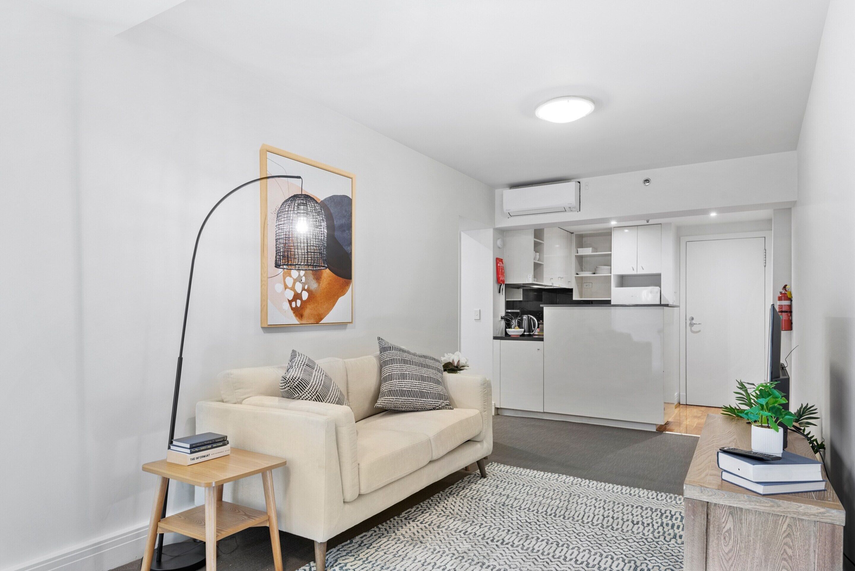 A modern living room with a sofa, a small side table, and a floor lamp. There is a kitchen area with cabinets and a countertop visible through an open door.