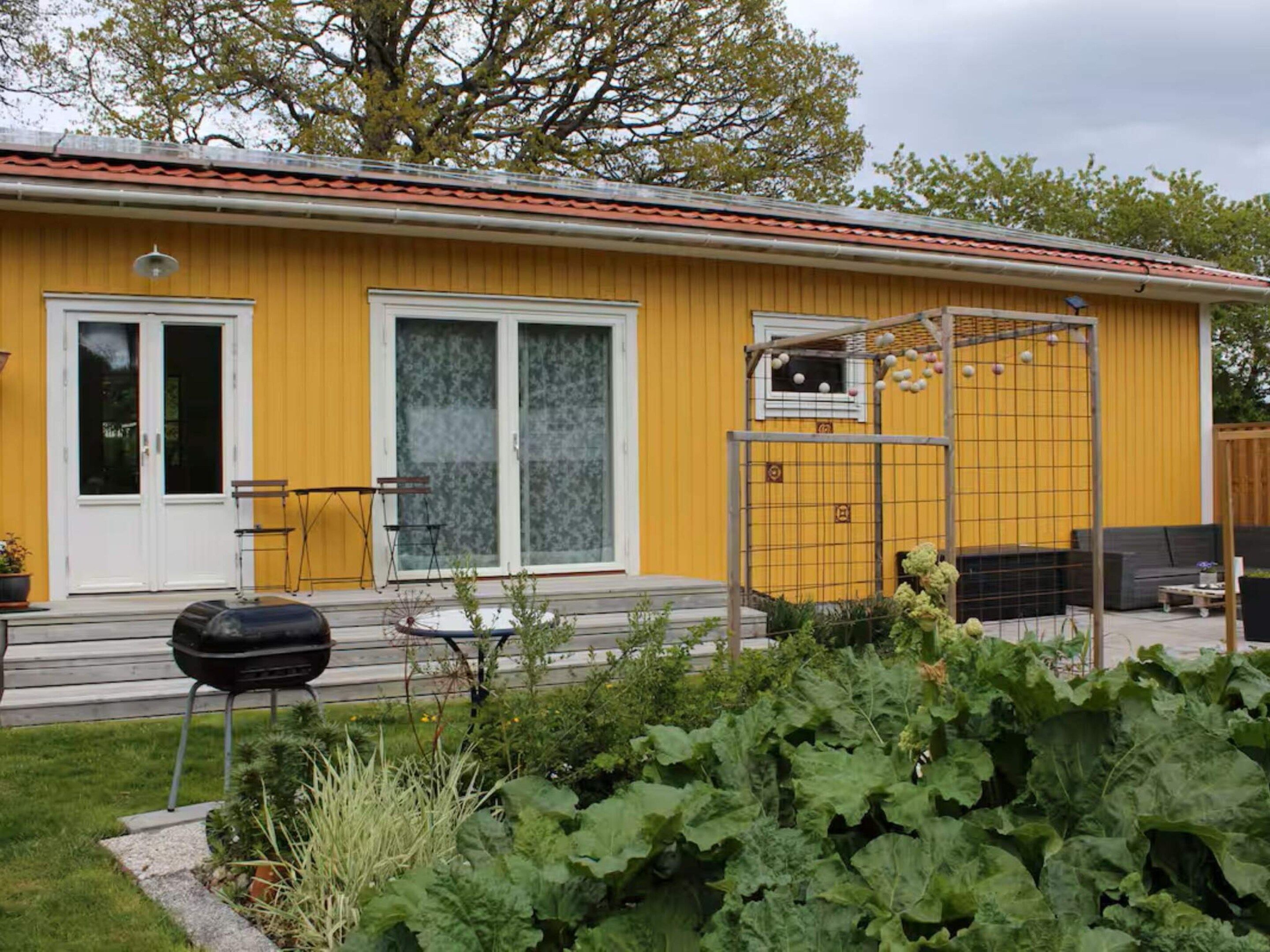 Een geel gebouw met een rood dak, een schuifdeur, een klein raam en een tuin met planten.