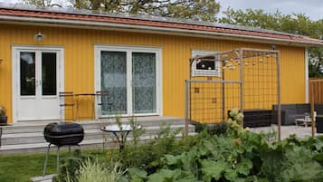 Edifício amarelo com telhado vermelho, porta de correr de vidro, uma pequena janela e área de jardim com plantas.