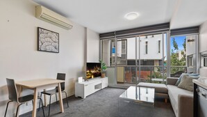 A modern living room with a dining table, sofa, and a TV. There is a balcony with a view of buildings and trees. - Fitzroy North 1-bed With Balcony by Edinburgh Park (Melbourne)