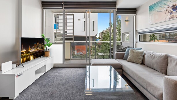 A modern living room with a large flat-screen TV, a sofa, a coffee table, and a balcony with a city view. - Fitzroy North 1-bed With Balcony by Edinburgh Park (Melbourne)