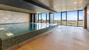 A modern indoor pool with a tiled edge, large windows offering a city view, and a wooden ceiling. - Modern 1-bed in Hip Southbank With Pool & Gym (Melbourne)