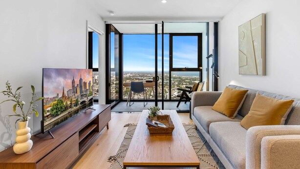 A modern living room with a large TV, a sofa, a coffee table, and a view of a cityscape through large windows. - Modern 1-bed in Hip Southbank With Pool & Gym (Melbourne)