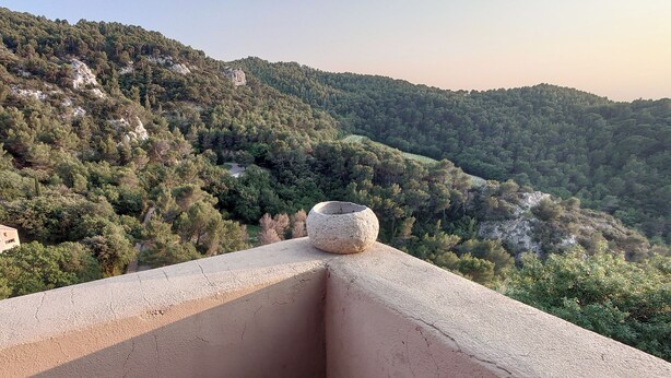 Un balcone con un vaso in pietra, affacciato su un pendio ricoperto da una fitta boscaglia.