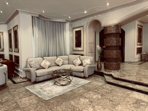 Een ruime woonkamer met een beige hoekbank, een glazen salontafel en een kamerkleed met een patroon.