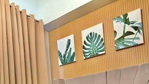 Un lit bien fait, recouvert d’une couverture grise, avec une serviette blanche et des coussins décoratifs, dans une chambre moderne aux murs lambrés et ornée de deux tableaux représentant des plantes.