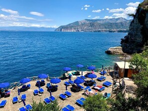 A beach with blue umbrellas and sun loungers, a rocky cliff, and a view of the sea and mountains in the background. - Gaiano APT Casette (Sorrento)