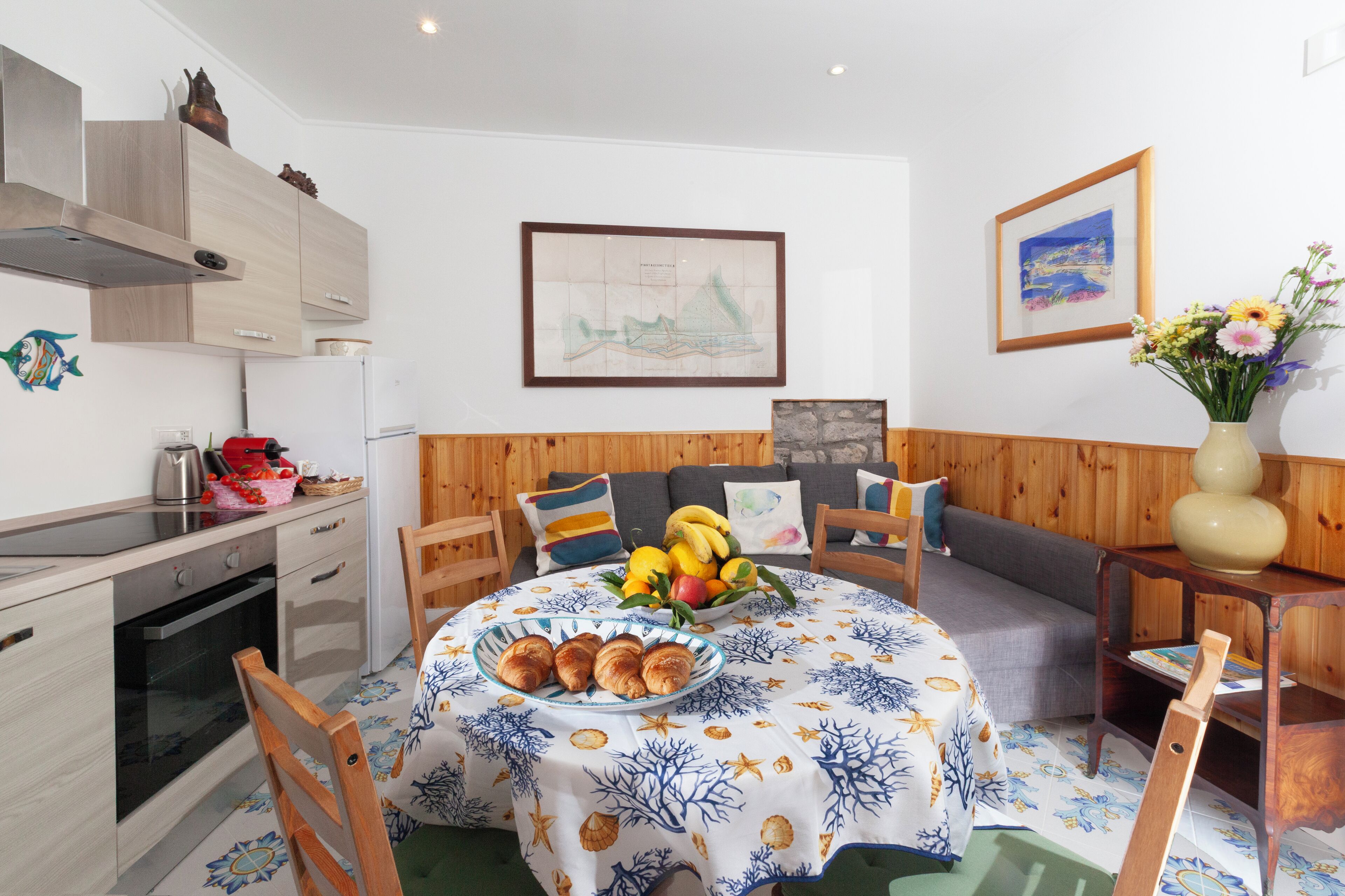 Un comedor con una mesa dispuesta para el desayuno, una cocina con armarios de madera y una sala de estar con sofá y cojines.