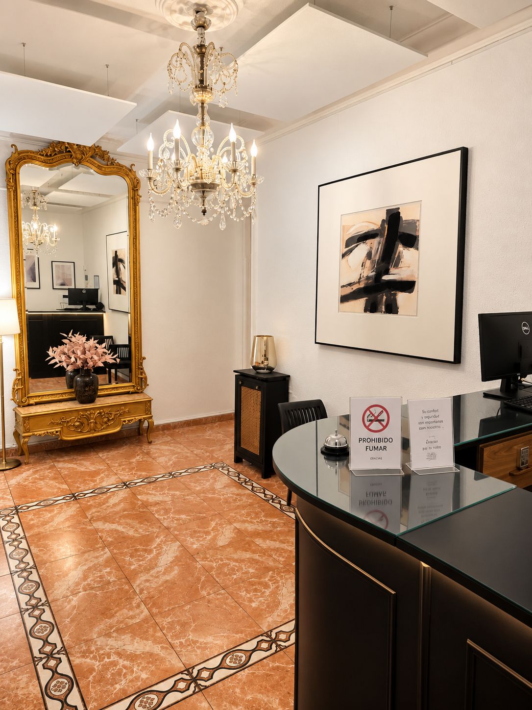 Un hall d’accueil d’hôtel avec un grand miroir, un lustre, un tableau abstrait encadré et un comptoir en verre sur lequel est apposé un panneau « interdiction de fumer ».