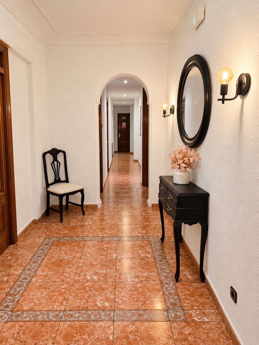 Un pasillo con una silla, una consola con un florero y un espejo montado en la pared.