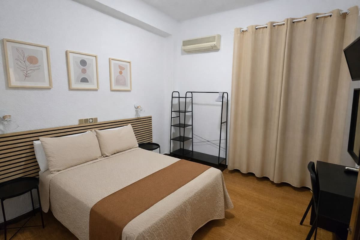 A bedroom with a bed, a desk, a chair, a shelf unit, and three framed artworks on the wall.