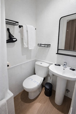 Un baño pequeño y moderno con inodoro, lavamanos y espejo. Hay un perchero sobre el inodoro y un cubo de basura junto al lavamanos.