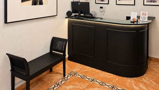 A reception area with a black desk, a bench, a chandelier, and framed artwork on the walls. - Hostal Prada (Madrid)