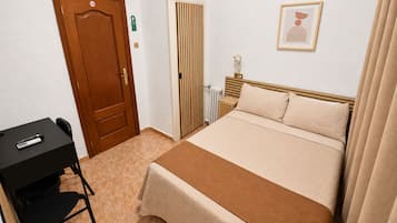 A hotel room with a bed, a desk with a chair, a radiator, and a framed picture on the wall.