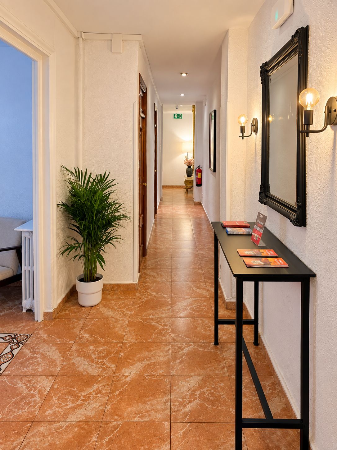Pasillo de hotel con una planta en maceta, una mesa con folletos y un espejo con apliques de pared.