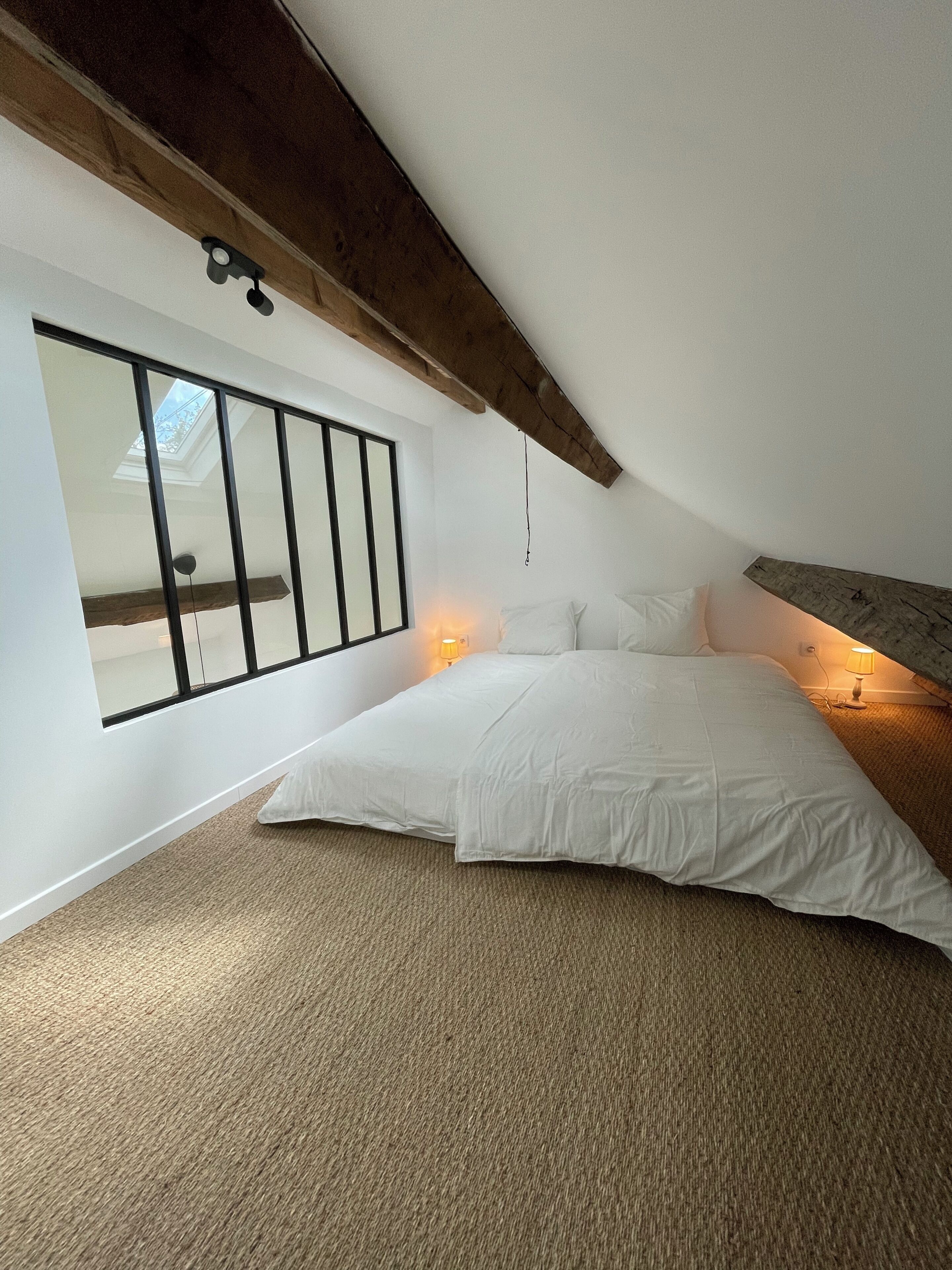 Une chambre avec un plafond en pente, une grande fenêtre, un lit avec une literie blanche et une lampe sur la table de chevet.