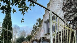 Une cour dallée de pierres, avec un bâtiment à l’aspect rustique, un portail en métal et un grand arbre.