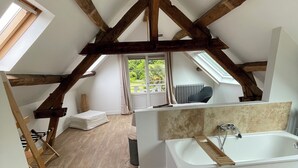 Une chambre moderne aménagée dans les combles, avec un plafond en pente, des poutres en bois, un puits de lumière, une grande fenêtre, une baignoire et une échelle en bois.