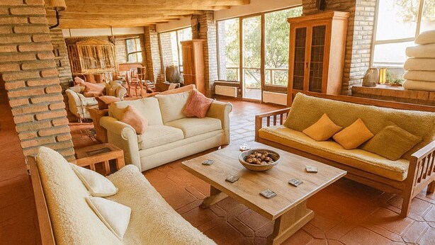 Un amplio salón con muebles de madera, chimenea de piedra y grandes ventanales que ofrecen vistas al exterior.