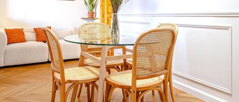 A dining area with a glass table surrounded by wicker chairs, a white sofa with orange cushions, and a potted plant.