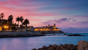 Un pueblo costero al atardecer con edificios históricos, una construcción fortificada y palmeras.