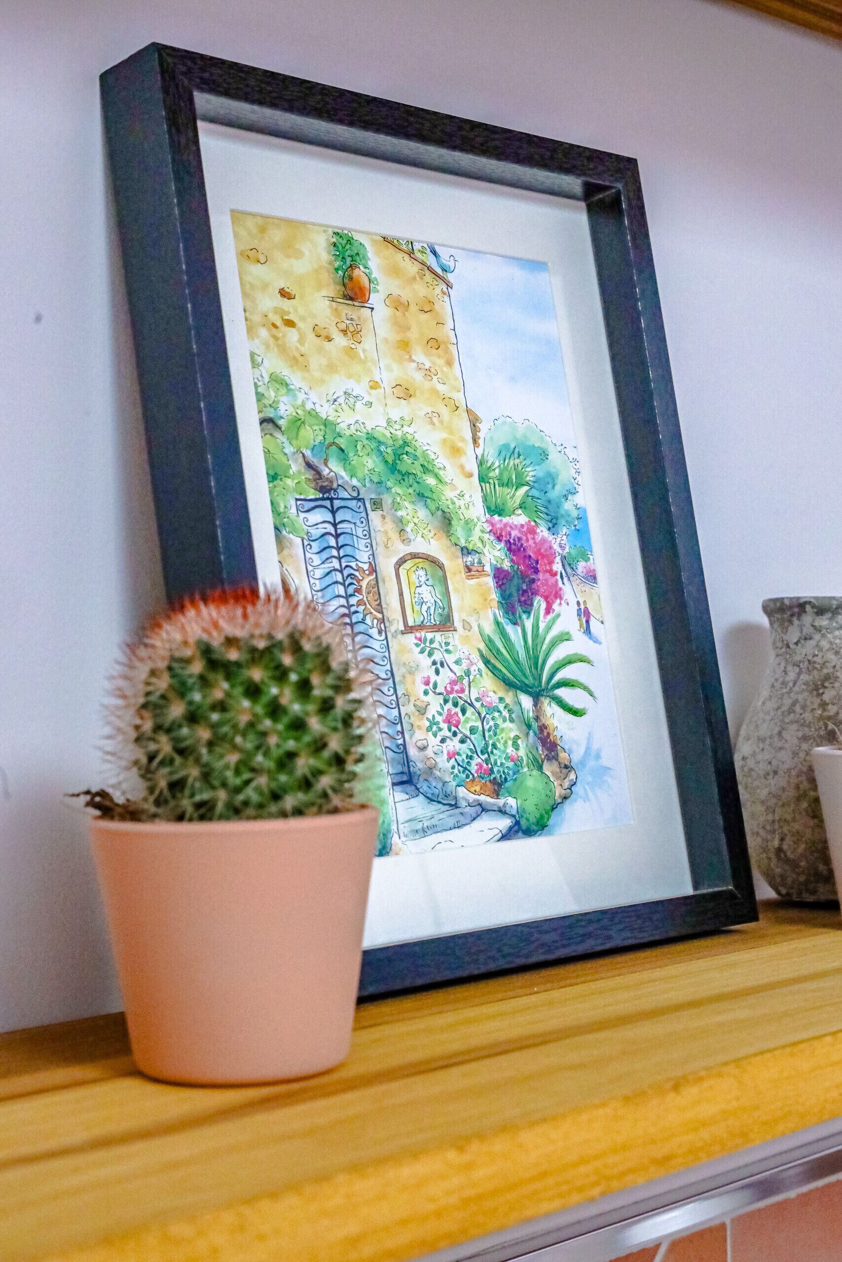Una acuarela enmarcada que muestra un jardín colorido con un cactus en una maceta rosa sobre un estante de madera.