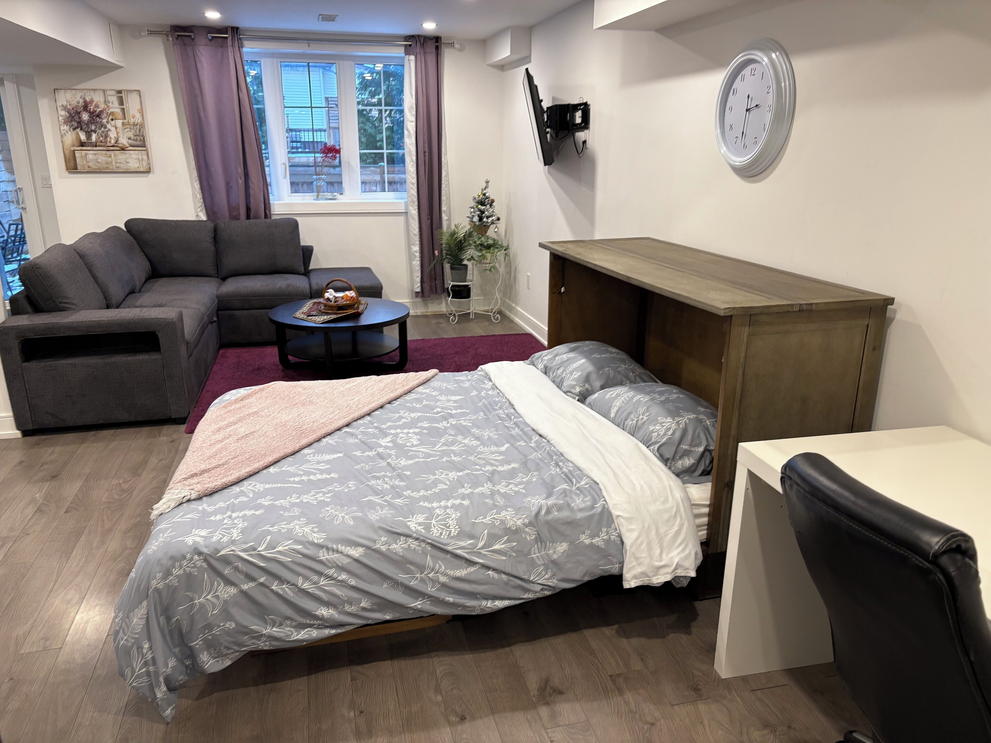 Un salon moderne avec un canapé, une table basse ronde, un lit avec une literie à motifs, un meuble en bois et une horloge au mur.