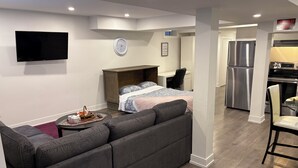 Un soggiorno moderno con un divano ad angolo grigio, una TV a schermo piatto montata a parete, un tavolino rotondo con un cesto e un letto con copripiumino fantasia.