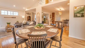 A spacious living room with a round wooden dining table, multiple chairs, and a central plant. There is a sofa, a coffee table, and a fireplace in the background. - Walk to Main St & Swamp Rabbit Trail (Travelers Rest)