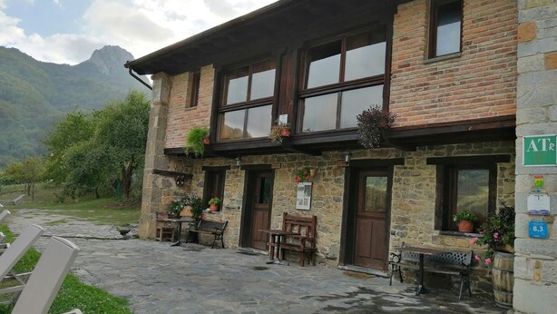 Un bâtiment en pierre avec une façade en bois, des places assises à l’extérieur et une vue sur les montagnes en arrière-plan.