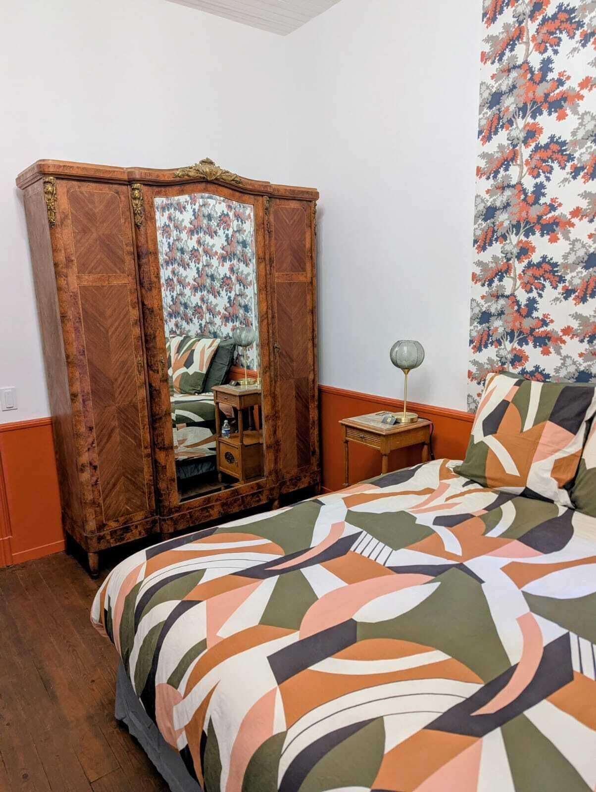 A bedroom with a bed, a wooden wardrobe, a small table, and a mirror.