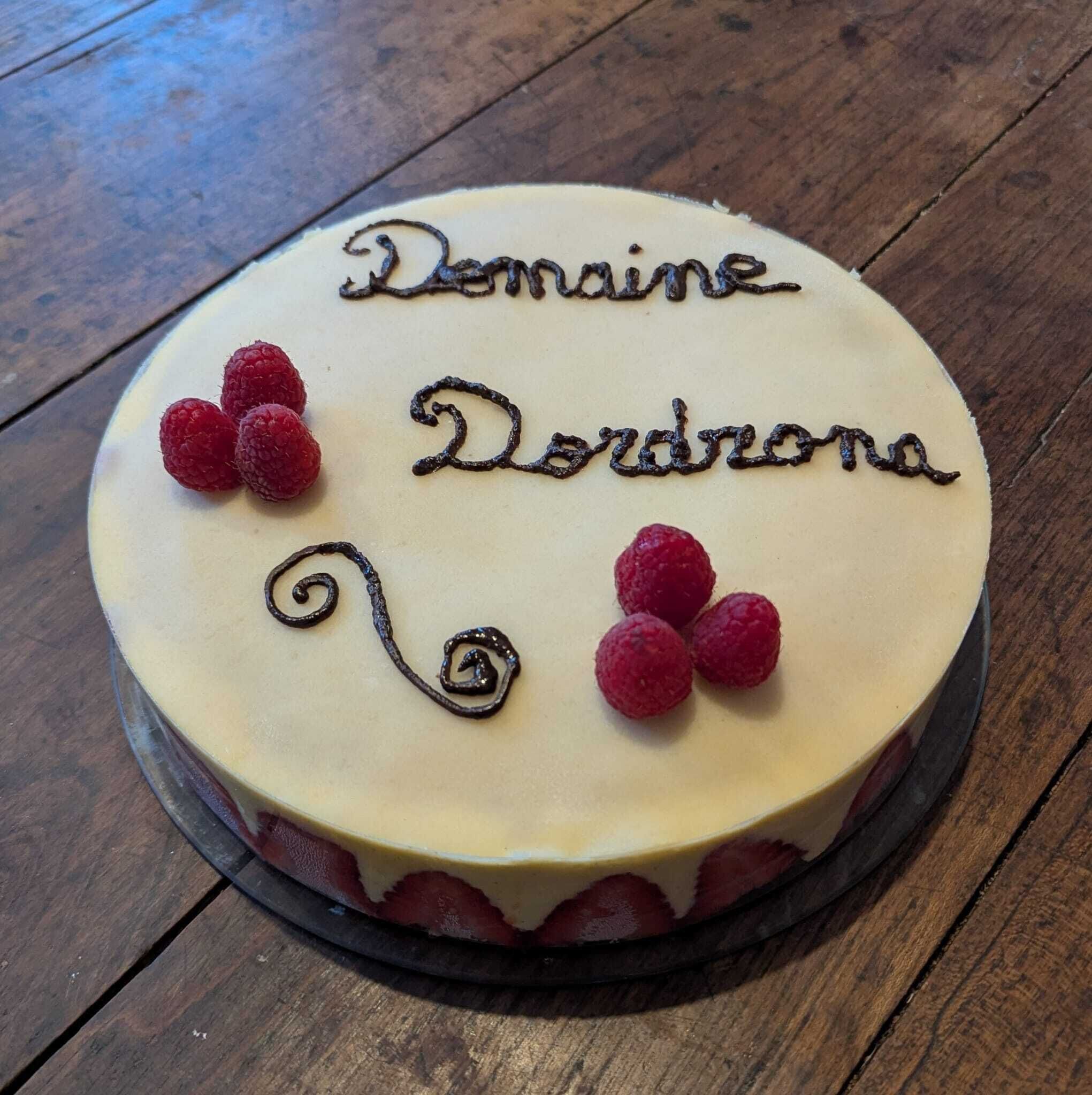 A round cake with 'Domaine Dordona' written on top, decorated with raspberries and chocolate swirls.