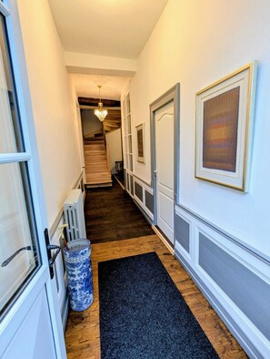A narrow hallway with a wooden floor, white walls, and a black carpet. A radiator and a blue and white vase are visible on the left. - Domaine de la Dordrona (Bourdeilles)