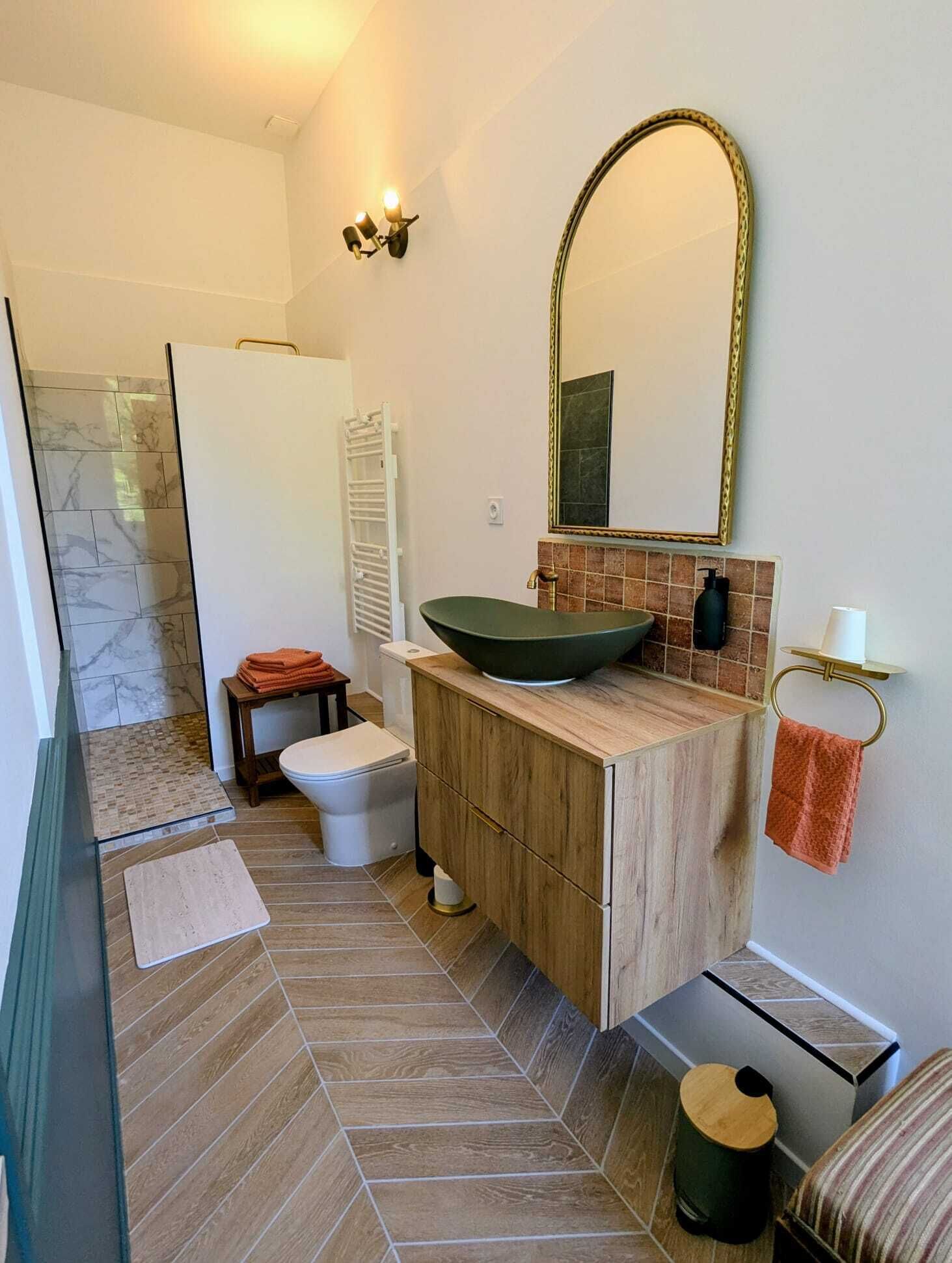 Un baño con ducha revestida de mármol, inodoro blanco, tocador de madera con lavabo verde y un espejo grande.
