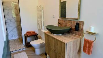 Un baño con ducha revestida de mármol, inodoro blanco, tocador de madera con lavabo verde y un espejo grande.