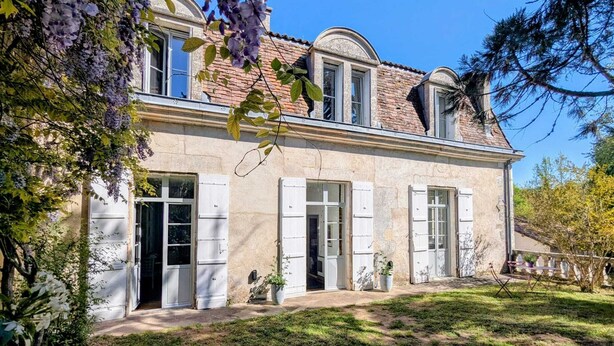 Une maison à deux étages avec une façade en briques, des volets blancs et un jardin où poussent différentes espèces de plantes.