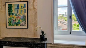 Une pièce décorée d’un tableau encadré représentant un paysage coloré, une fenêtre donnant sur des immeubles et une petite table sur laquelle est posée une plante en pot.