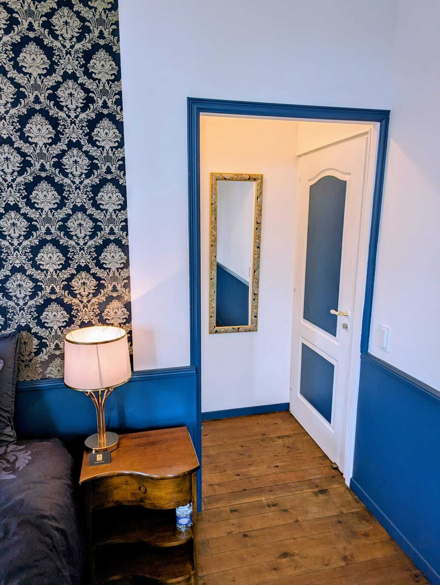Una habitación con pared azul, papel tapiz estampado, cama, mesita de noche con lámpara y puerta con espejo.
