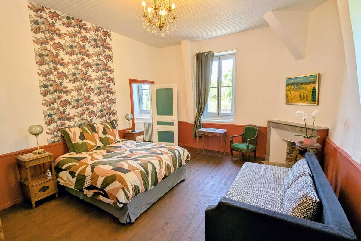 Een slaapkamer met een bed, een stoel, een tafel, een open haard en een raam.