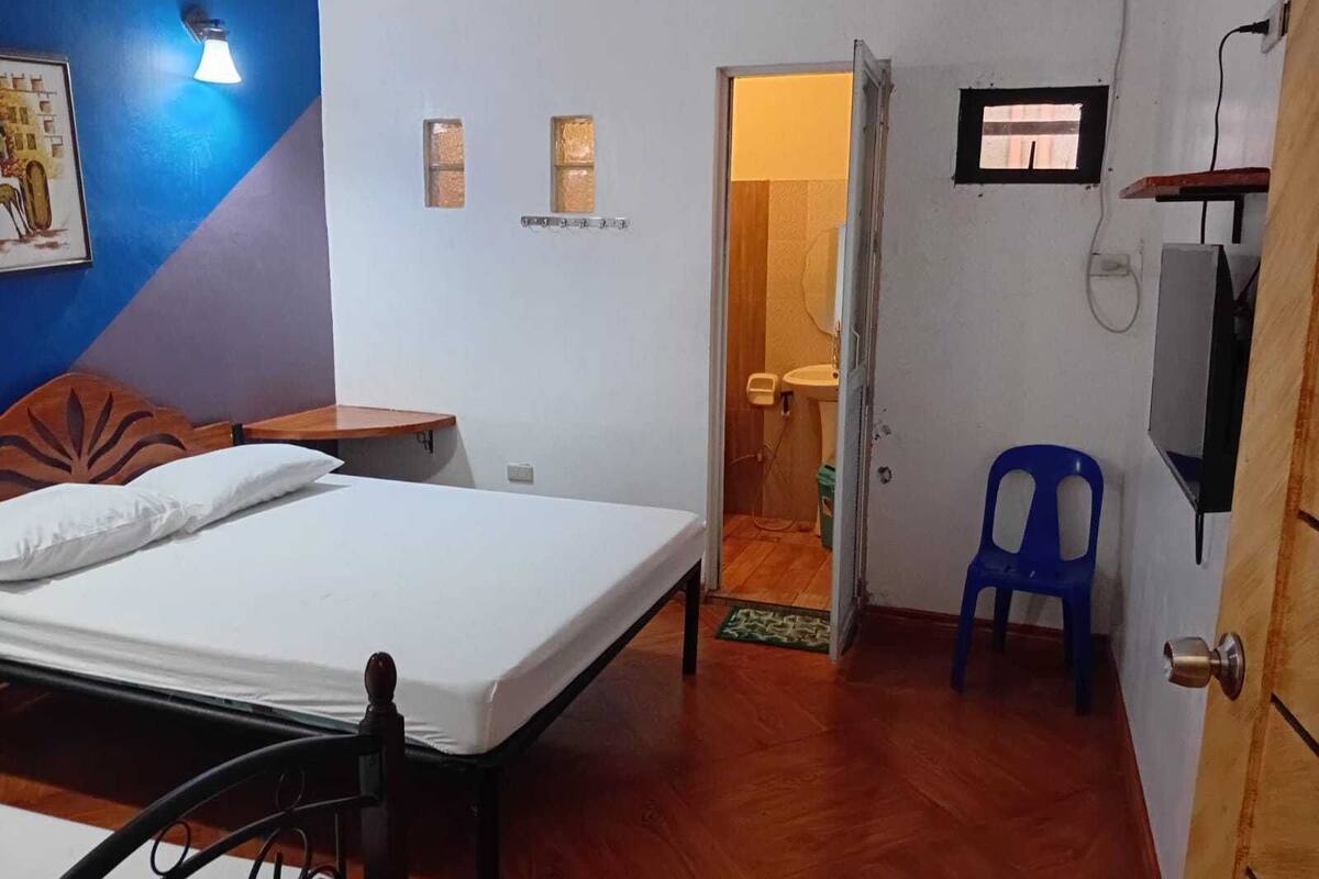 Une chambre à coucher avec un lit, une tête de lit en bois, une table de chevet, une chaise bleue et une salle de bain visible à travers une porte ouverte.