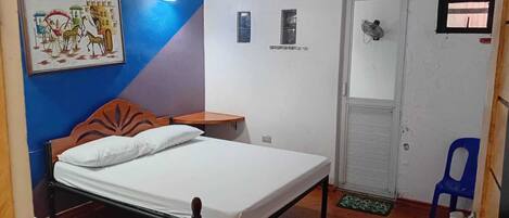 A bedroom with a bed, a nightstand, a wall-mounted light, a door, a small window, and a blue chair.