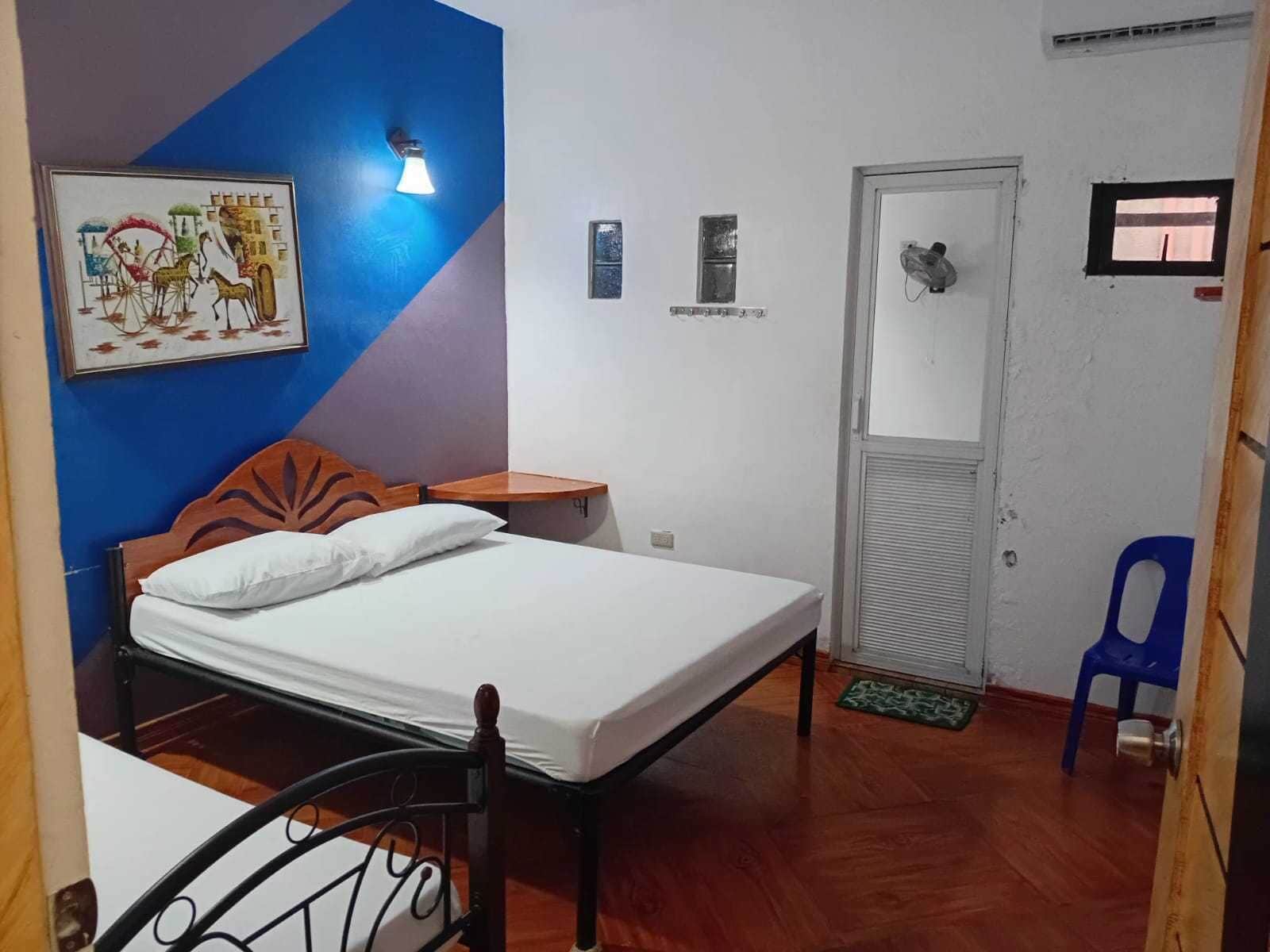 A bedroom with a bed, a nightstand, a wall-mounted light, a door, a small window, and a blue chair.