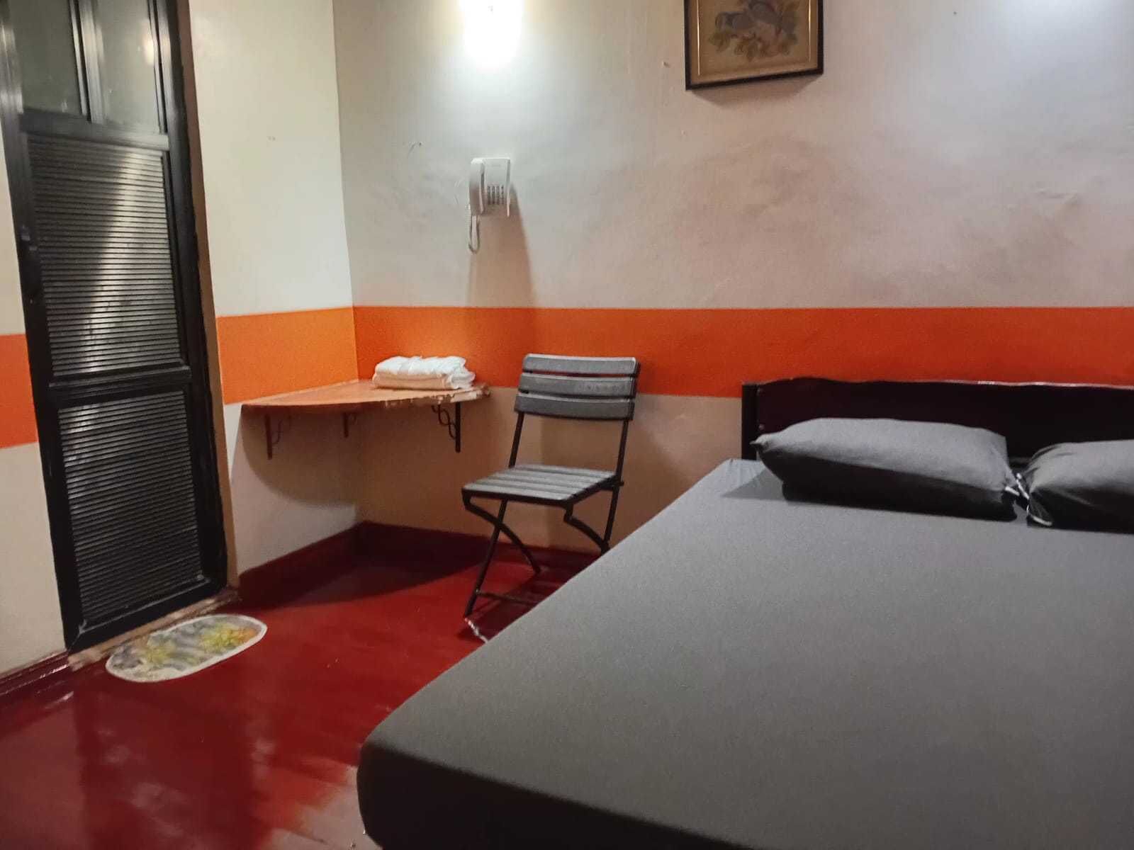 A hotel room with a bed, a chair, a small table with a towel, a wall-mounted phone, and a door with a metal handle.