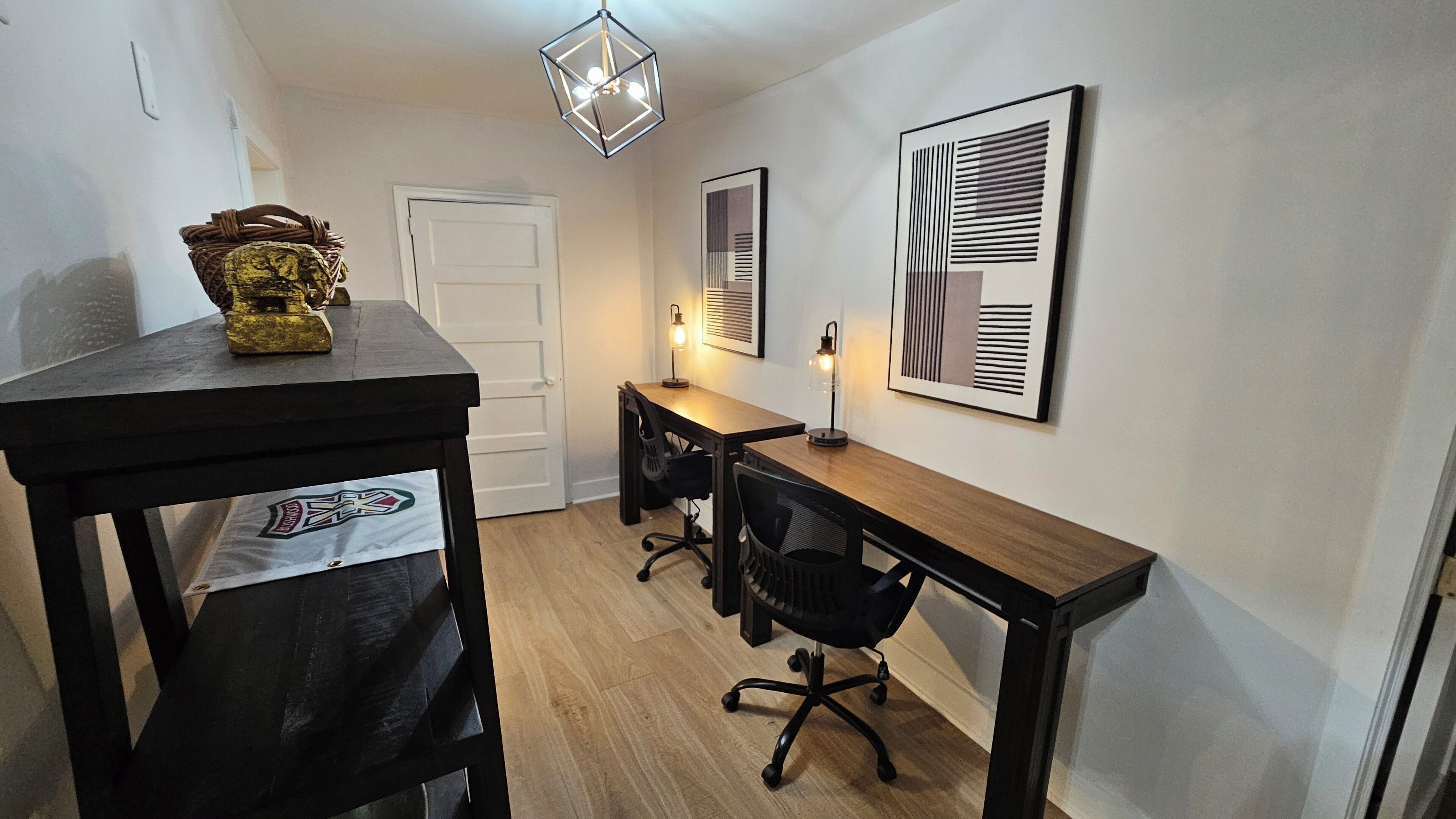 Une petite pièce bien éclairée, avec un bureau en bois, deux chaises de bureau noires, un vase décoratif et deux œuvres d’art abstraites encadrées accrochées au mur.