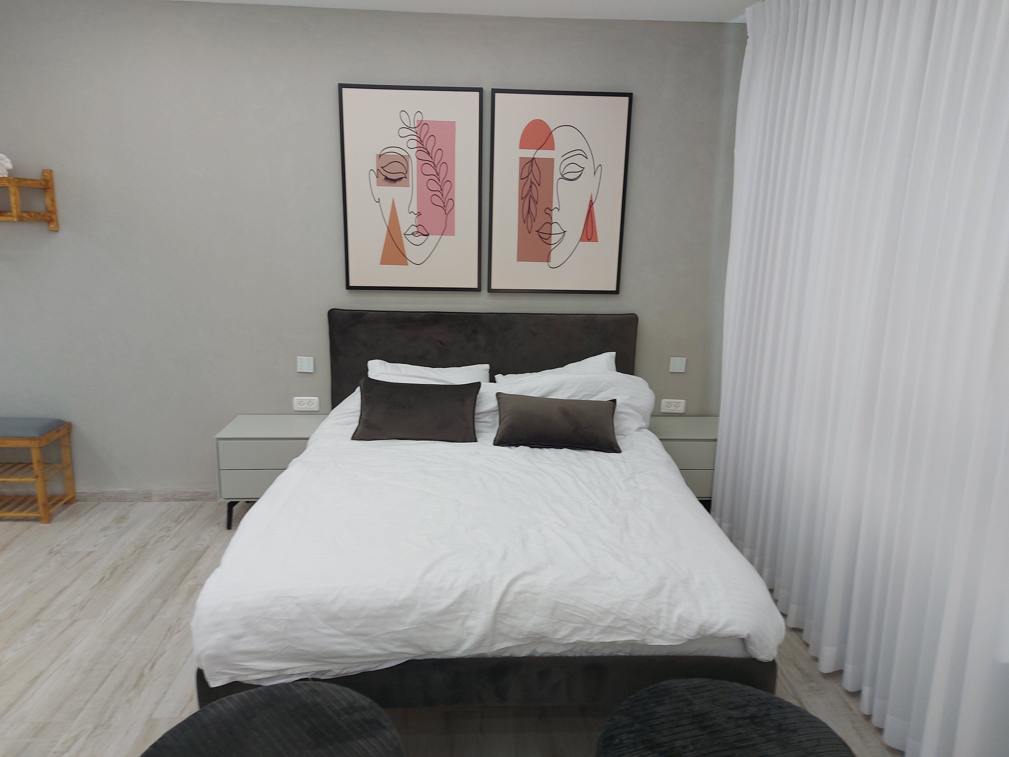 Un dormitorio con una cama, dos mesitas de noche, una silla de madera y dos cuadros enmarcados en la pared.