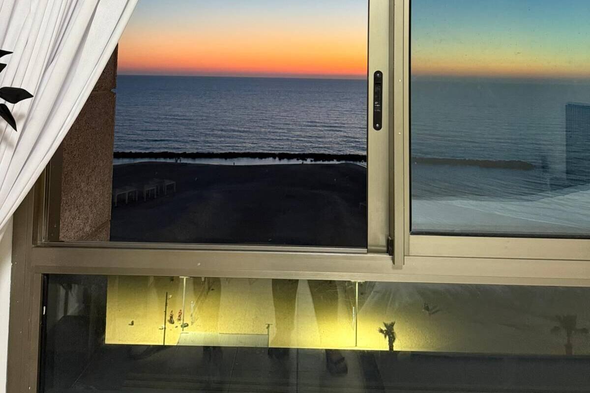 Une chambre avec vue sur l’océan au coucher du soleil, une porte-fenêtre coulissante et une fenêtre donnant sur la plage.